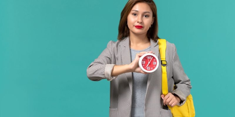 front-view-female-student-grey-jacket-wearing-her-yellow-backpack-holding-clocks-light-blue-wall