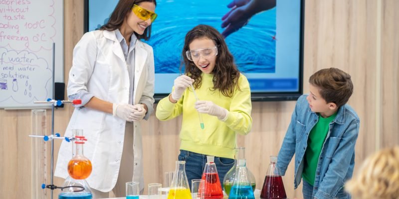 Good result. Rejoicing girl looking at test tube in hands and watching teacher with children in chemistry class